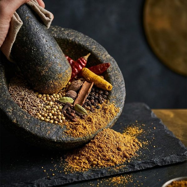Whole spices — cumin, coriander seed, cardamom, cloves, cinnamon, dried chillies and turmeric — being ground in a stone pestle and mortar